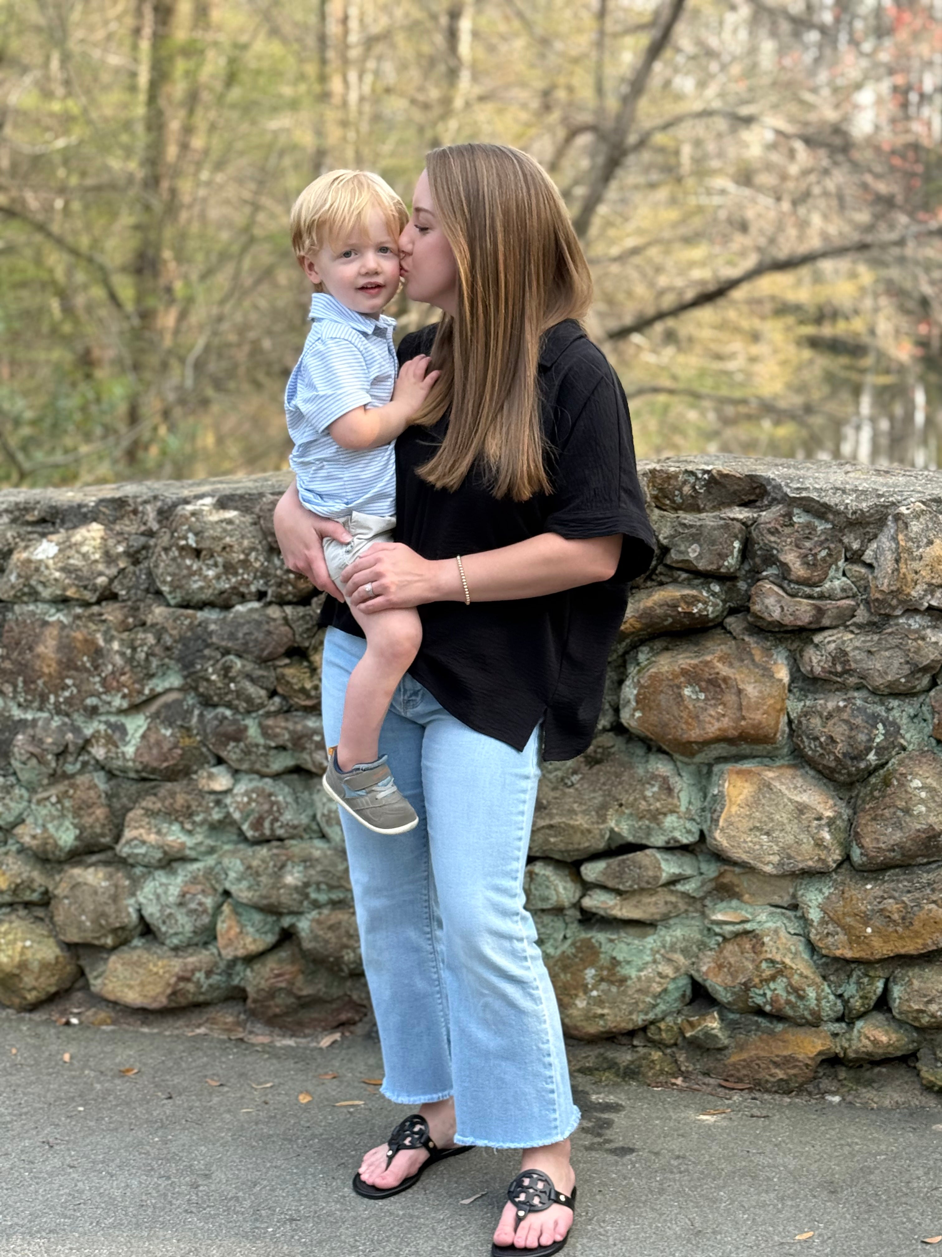 Woman holding a child and kissing them on a stone wall with trees in the background