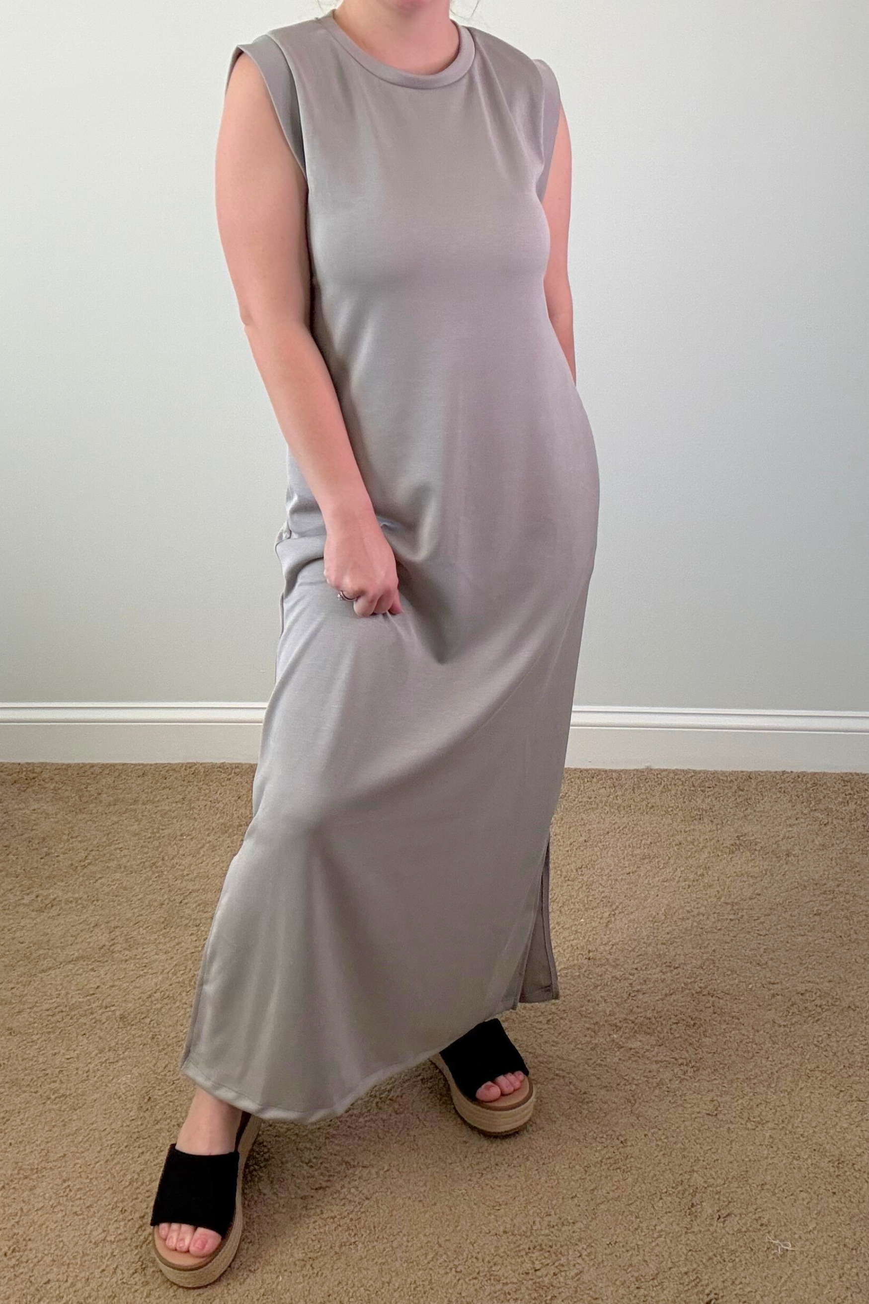 Person wearing a long light gray dress standing on a carpeted floor with a white wall background.
