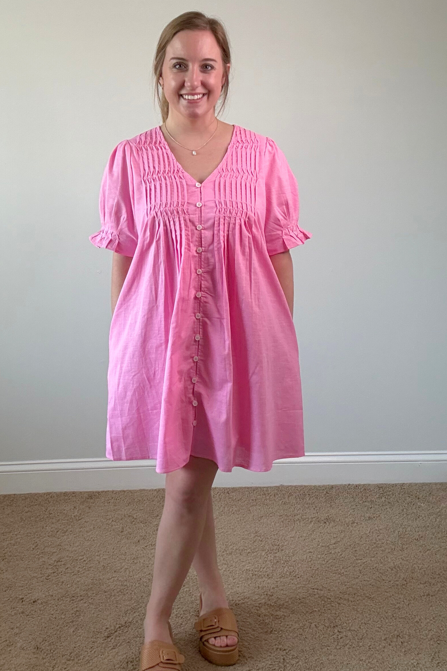 Woman wearing a pink dress standing against a plain wall.
