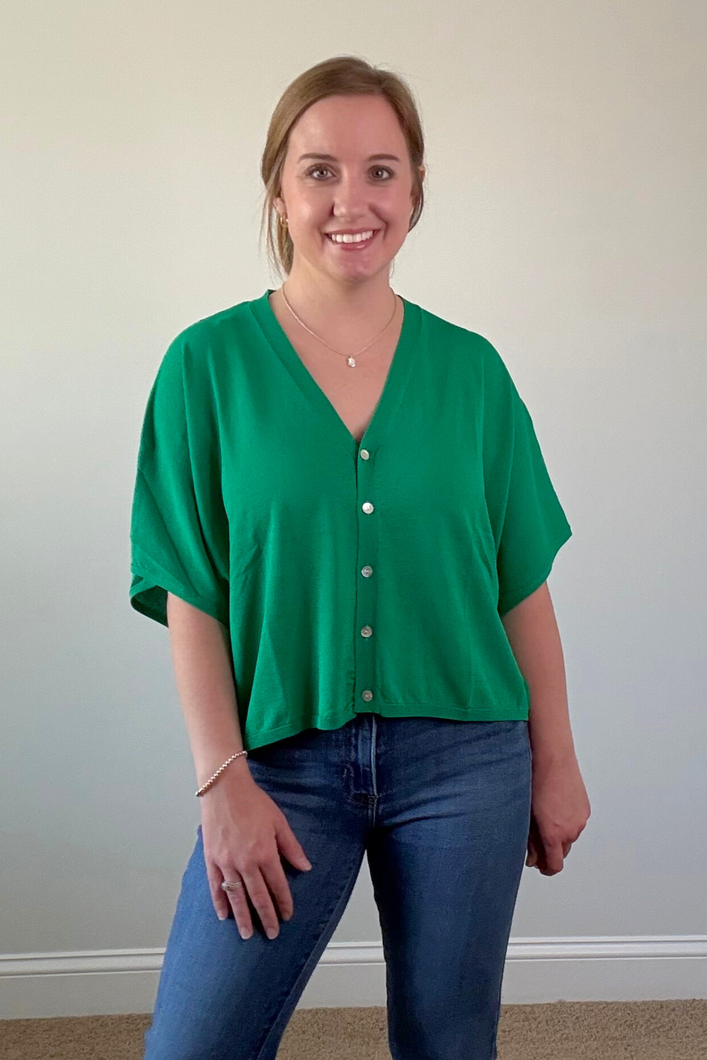 Woman wearing a green blouse with white buttons and blue jeans against a plain wall.