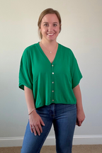 Woman wearing a green blouse with white buttons and blue jeans against a plain wall.