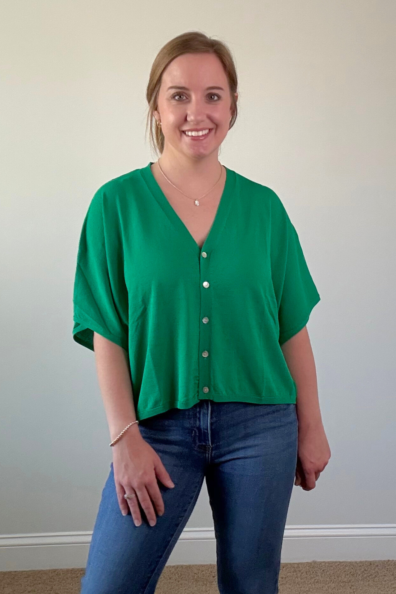 Woman wearing a green blouse with white buttons and blue jeans against a plain wall.