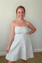 Woman wearing a strapless white dress with blue stripes against a plain wall.