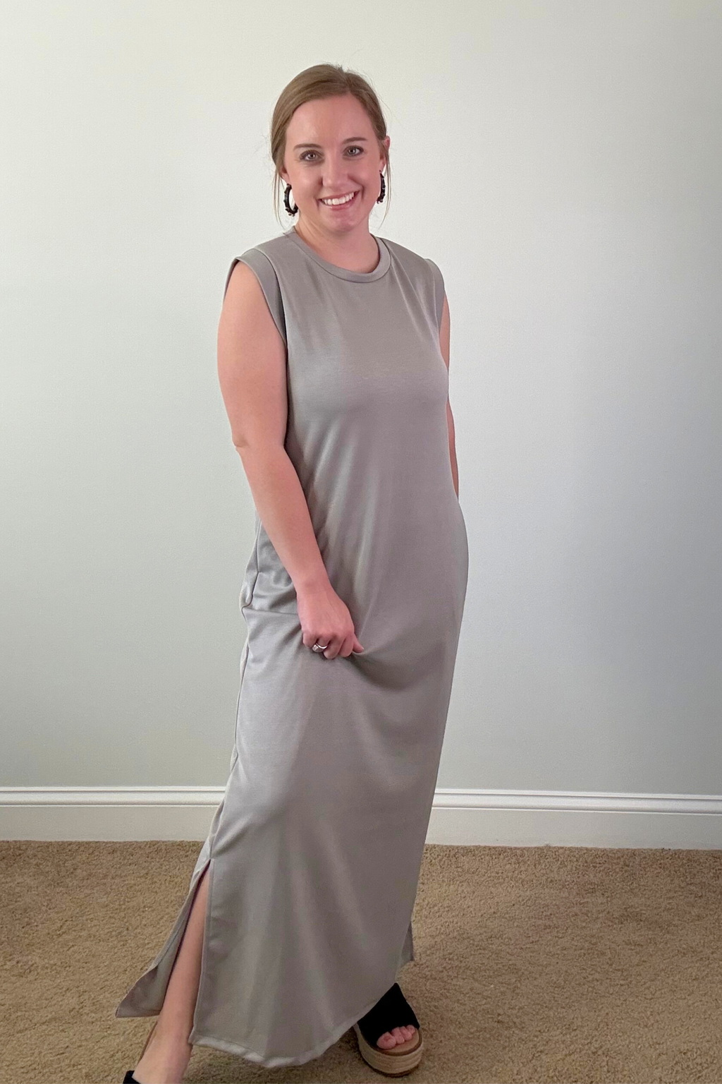 Woman wearing a gray sleeveless dress standing against a plain wall.